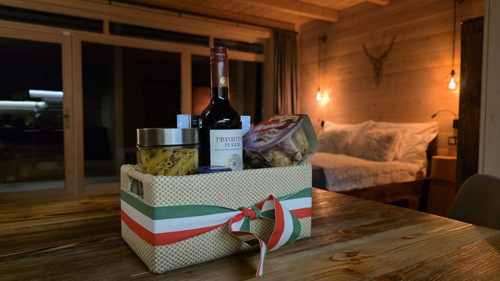 vacsora, olasz vacsora, A Chalet gerendaház asztalán egy kosárban olasz vacsorabekészítés látható tésztaétellel, desszerttel és borral, a kosár olasz zászló szalaggal van átkötve.