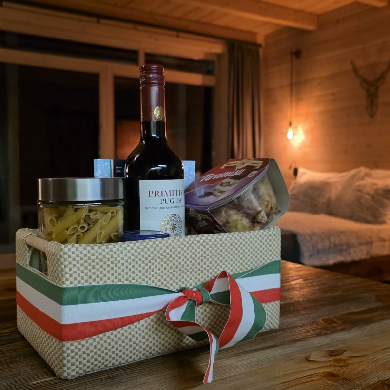 vacsora, olasz vacsora, A Chalet gerendaház asztalán egy kosárban olasz vacsorabekészítés látható tésztaétellel, desszerttel és borral, a kosár olasz zászló szalaggal van átkötve.